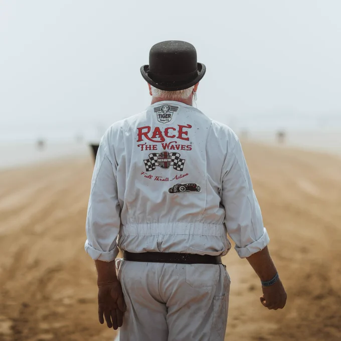 A view of the back of a man wearing a white outfit and black hat, with Race the Waves written across the back of his shirt