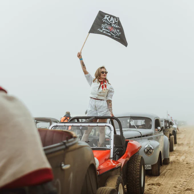 A woman wearing white and waving a Race the Waves flag from one of the vintage vehicles, which are parked in a line on Bridlington's south beach