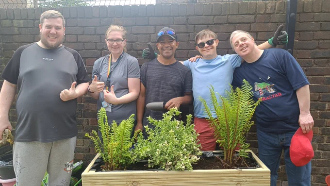 Three Share students and two members of staff posing behind a wooden planter filled with green plants. They are smiling and some of them have their thumbs up.