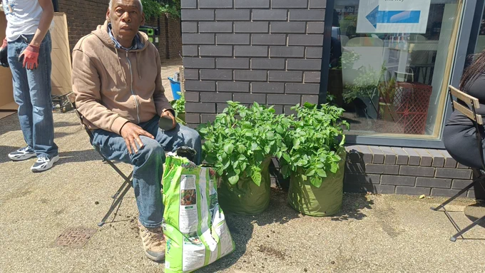 A Share student sat on a chair next to two potato plants in bags. He is holding a trowel.