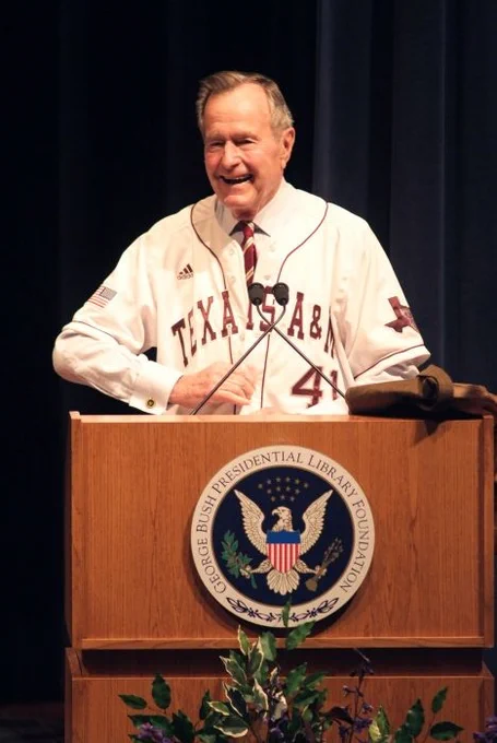 Former President George H.W. Bush speaking at a podium bearing the seal of the George Bush Presidential Library and Museum, wearing a Texas A&M baseball jersey.