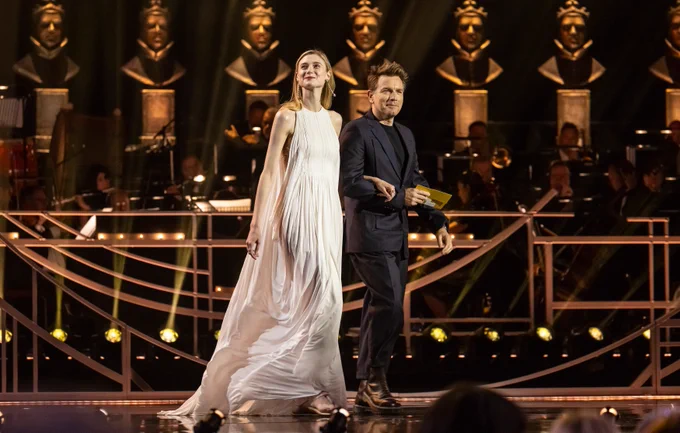 Elizabeth Debicki and Ewan McGregor walk onstage at the Olivier Awards. Elizabeth wears a long white dress with blonde hair and Ewan wears a dark suit with short dark hair.