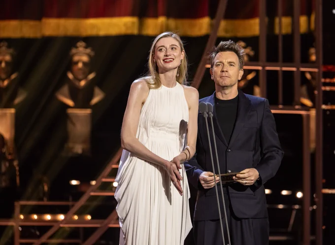 Elizabeth Debicki and Ewan McGregor present onstage at the Olivier Awards. Elizabeth wears a long white dress with blonde hair and Ewan wears a dark suit with short dark hair.