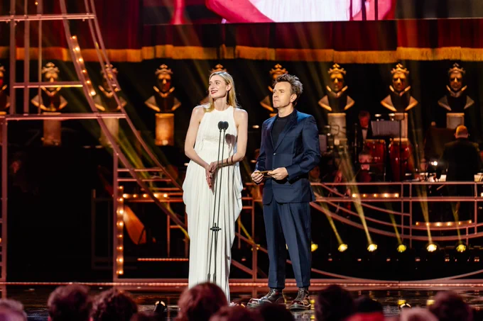 Elizabeth Debicki and Ewan McGregor present onstage at the Olivier Awards. Elizabeth wears a long white dress with blonde hair and Ewan wears a dark suit with short dark hair.