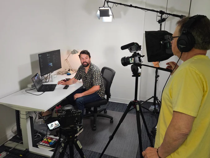 A man in foreground stands behind a camera and further filming equipment pointing at a man sitting at a desk with laptop, screen, mouse, keyboard and lamp.