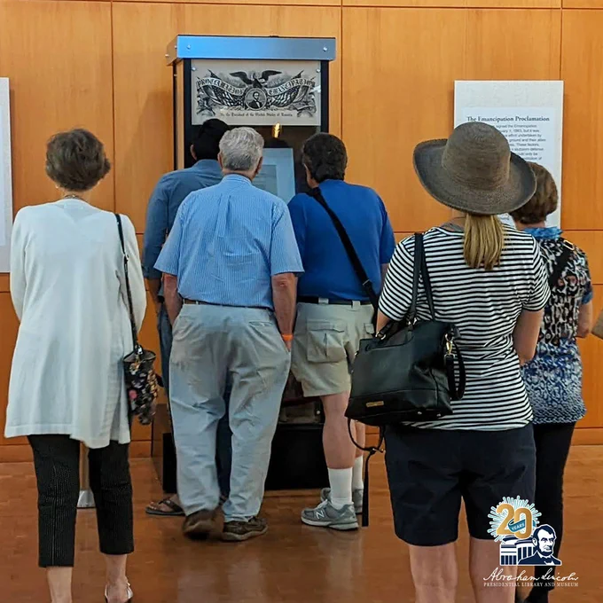 Visitors gather around a display case housing a rare copy of the Emancipation Proclamation at the Abraham Lincoln Presidential Library and Museum. The document is mounted upright with interpretive text nearby. A mix of museum guests, including seniors and a woman in a wide-brimmed hat, observe the exhibit. A 20th anniversary ALPLM logo appears in the lower corner.