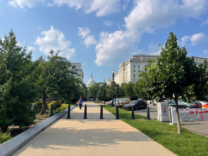 A paved sidewalk with trees and bollards leads toward the U.S. Capitol building under a partly cloudy sky, with nearby buildings and parked cars.