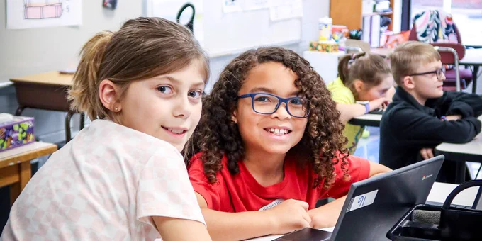 Two children smiling while using a laptop in a classroom setting.