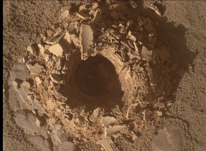 This is a close-up image of a drill hole in a flat, dusty, light-brown rock surface on Mars. The hole, made by the Curiosity rover, is surrounded by scattered, fractured rock fragments. The terrain is cracked and finely textured, piling up around the hole.