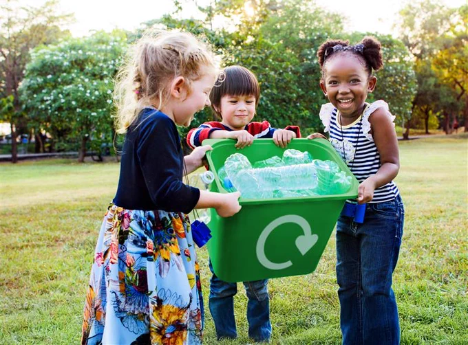 Three kids in a field holding a full recycling bin