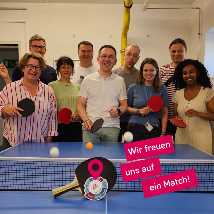 Foto einer Tischtennisplatte und Mitarbeitenden des BiB. Am Netz angelehnt liegt ein Tischtennisschläger, rechts daneben steht mit weißer Schrift auf pinkem Grund: "Wir freuen uns auf ein Match!"