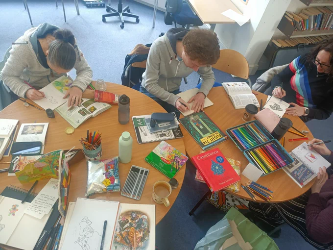Photo of a group of adults attending an art club at a library. The photo is taken from a height and shows adults sitting around library tables working on their illustrations inspired by various library books.