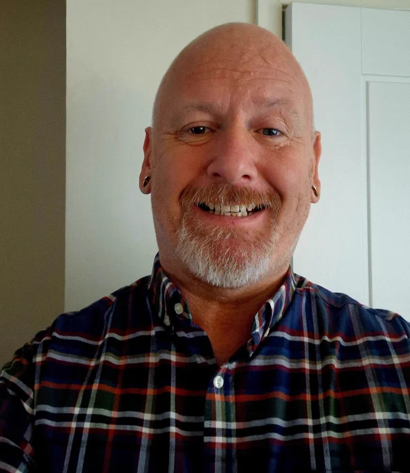 A selfie of Ian wearing a plaid shirt. The background includes a white door and part of a wall. 