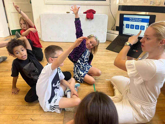 A group of young people sit on the floor with their hands up as the session leader points to an image on a laptop screen