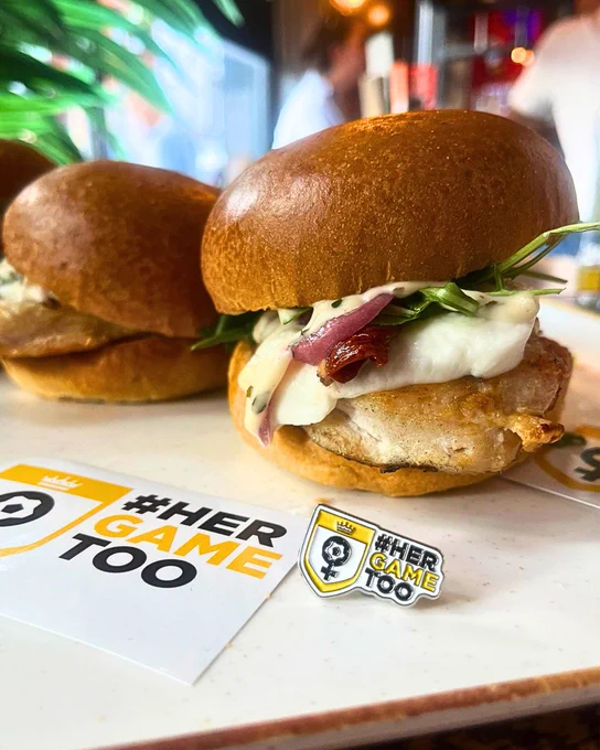 Close up image of Russo chicken burger with Her Game Too enamel pin badge and sticker in the foreground