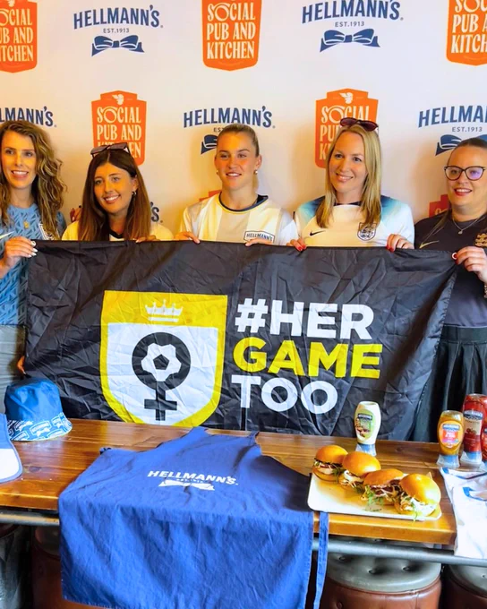 Her Game Too team members with Alessia Russo holding Her Game Too flag. On the table are 4 chicken burger, sauces, a Hellman's apron and Hellman's bucket hat. The backdrop contains Social Pub & Kitchen and Hellman's logos.