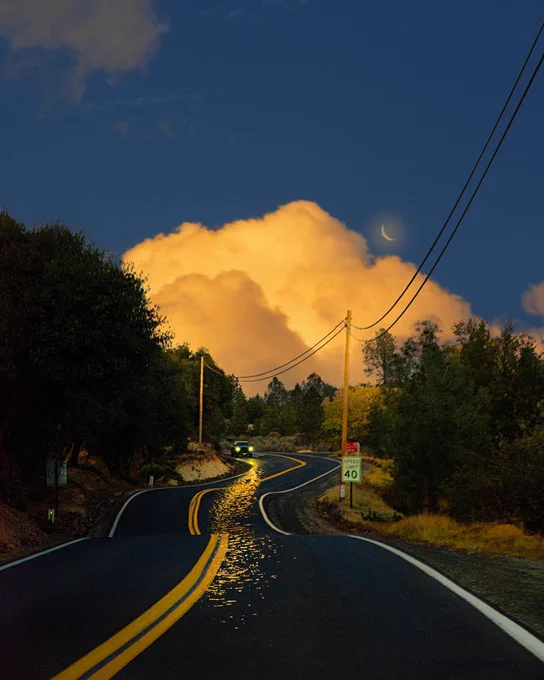 A curvy road at sunset with golden light on the pavement, glowing clouds, and a crescent moon in the sky.