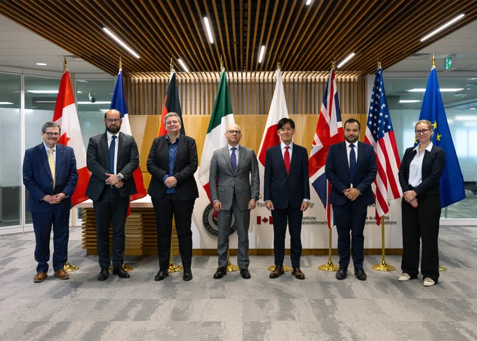 Members of the G7 cyber security working group in business attire stand in a row in front of flags from Canada, France, Germany, Italy, Japan, the UK, the USA, and the EU in a modern conference room. 