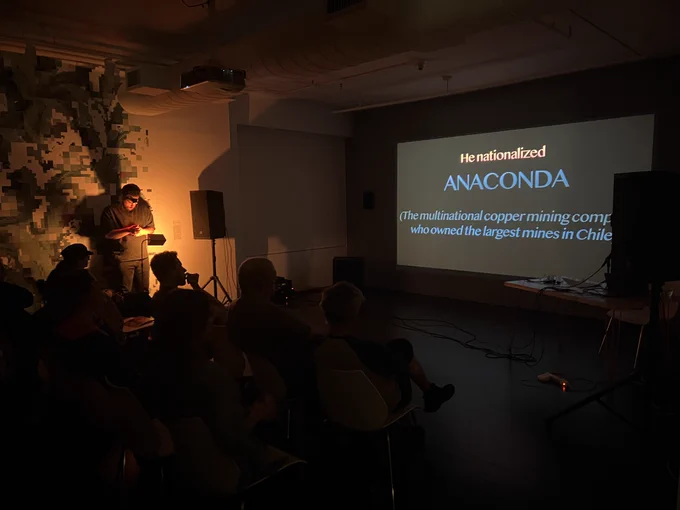 A performance in a dark gallery space. A performer is on the left is wrapping his arms in copper. On the right, a large projection has large text reading: “he nationalized Anaconda”. Audience members are in the dark.
