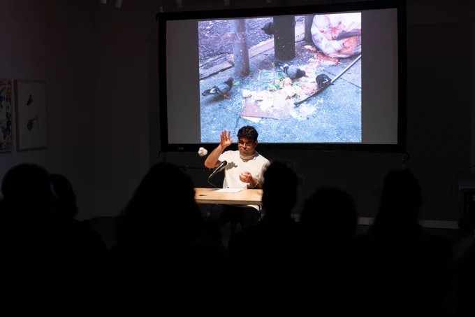 In a dark space, a spotlight is on a performer who is throwing a crumpled piece of paper onto the floor in front. Behind him, a large projection screen shows an image of trash on the sidewalk.
