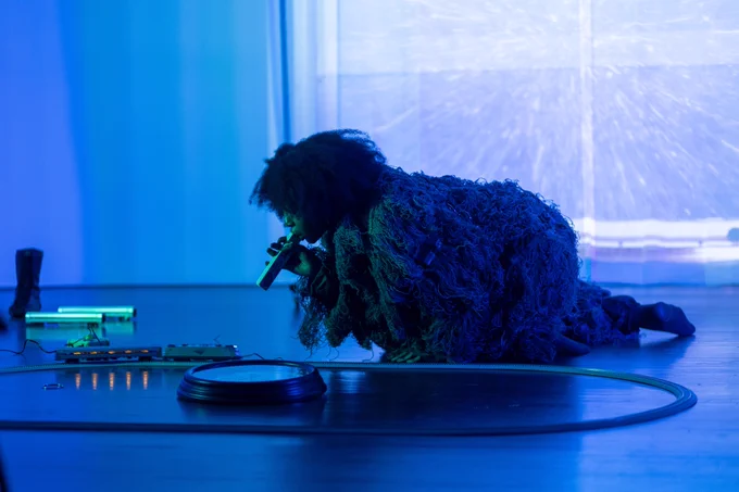 In a gallery space flooded in light blue light, a performer in a ghillie suit lays on the floor, tooting a horn at a model train set on front of them.
