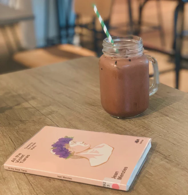 A photograph of a pink book with a Greek bust crowned with purple flowers on the cover: ‘All The Violet Tiaras’ by Jean Menzies lying on a table beside a mug of iced chocolate.