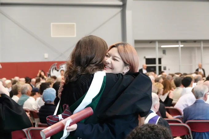 A graduating student holding a diploma and alumni tie is smiling while hugging another person.