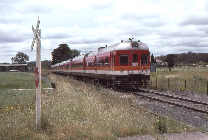 A 7-car DEB set diesel multiple unit train in Candy livery running on a single track line in a rural location approaches a level crossing marked with a set of crossbucks and a stop sign.