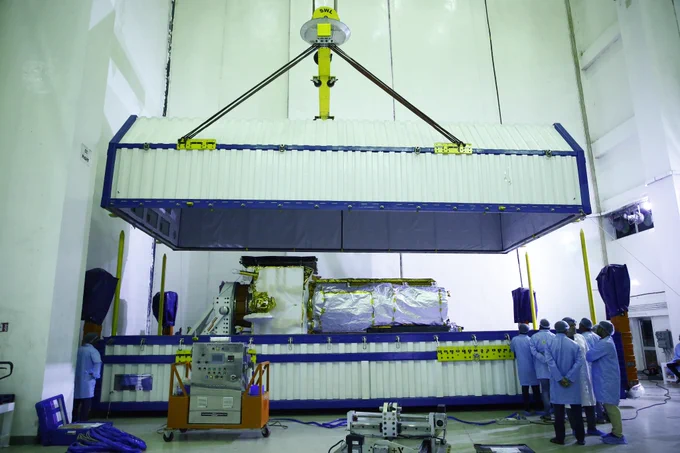 The lid of a specialized container is lifted to reveal NISAR, the Earth-observing radar satellite jointly developed by NASA and the Indian Space Research Organisation (ISRO), shortly after the spacecraft’s arrival at ISRO’s Satish Dhawan Space Centre in Sriharikota on India’s southeastern coast on May 16, 2025. The container is white and is being lifted above the spacecraft that is covered in silver blanketing, People in blue frocks stand to the right of the container. The image is slightly tinted in color because of fluorescent lights in the facility.
