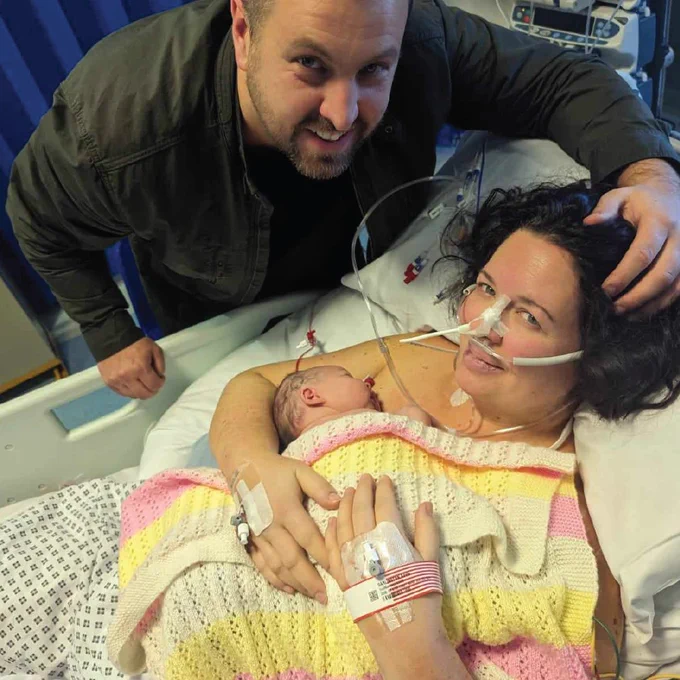 Adele lies in a hospital bed holding her newborn baby Jasmine against her chest, wrapped in a pastel striped knitted blanket. She has nasal oxygen tubing and IV lines, while the father touches her head and smiles at the camera.