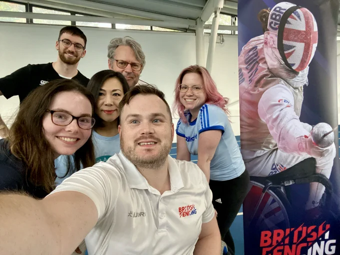 A selfie with six people smiling to the camera. They are next to a wheelchair fencing banner.