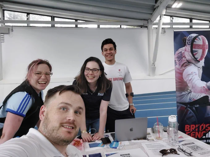 A selfie with four people smiling to the camera. They are next to a table with some things on it, and a wheelchair fencing banner is next to them.