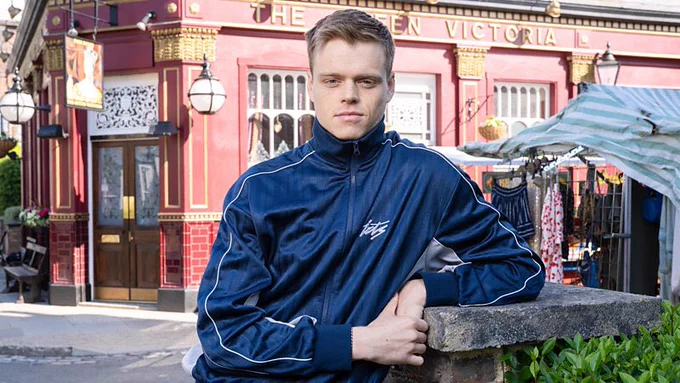 A young man in a tracksuit leans on a wall, against the backdrop of the Queen Victoria pub and market stalls