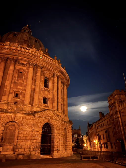 Night view of the Radcliffe Camera in Oxford under a starry sky with a bright moon illuminating the historic building and adjacent street.