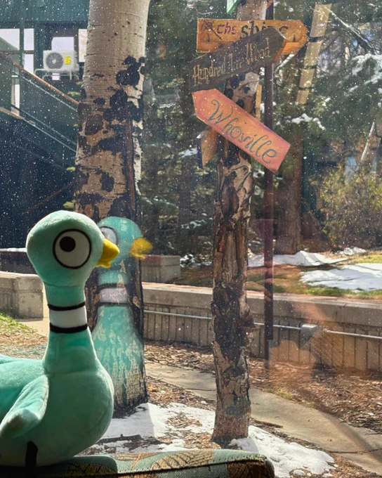 A plush toy Pigeon sits inside looking out a window at a snowy scene with directional wooden signs pointing to "The Shire" and "Whoville" attached to a tree.