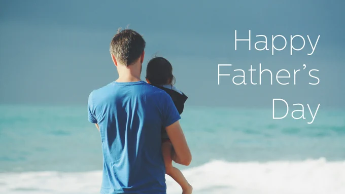 An individual carrying a child while standing on a beach, with text reading 'Happy Father’s Day' overlaying a blue sky.