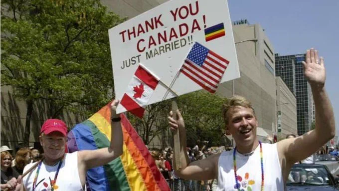 This photo captures a celebratory moment during what appears to be a pride parade. Two individuals are prominently featured, both smiling and waving. They are holding a sign that reads "THANK YOU! CANADA! JUST MARRIED!" with small flags of Canada and the United States attached to it. One of them is also holding a rainbow flag, symbolizing pride and support for the LGBTQ  community. The background shows a cityscape with tall buildings and a crowd of people, indicating a public event. 