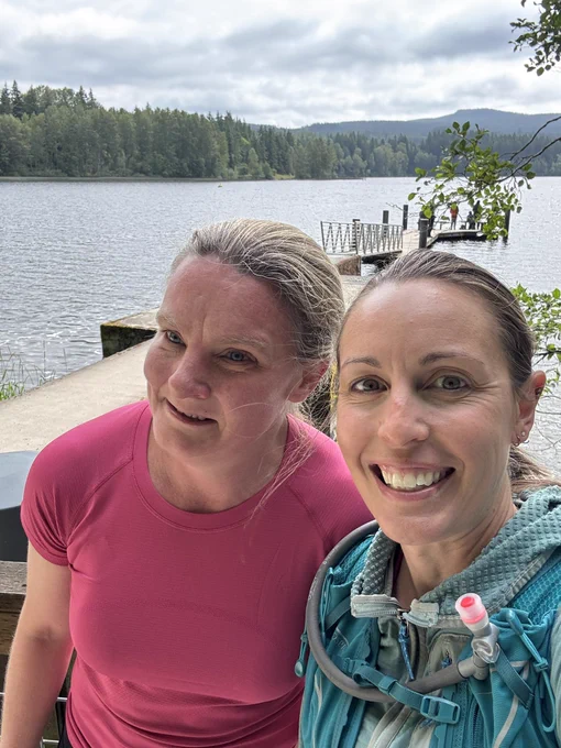Two women smiling for a selfie at Lake Padden. One is in a pink shirt, the other in a blue jacket with a hydration pack. The lake, a dock, and forested hills are in the background under a cloudy sky.