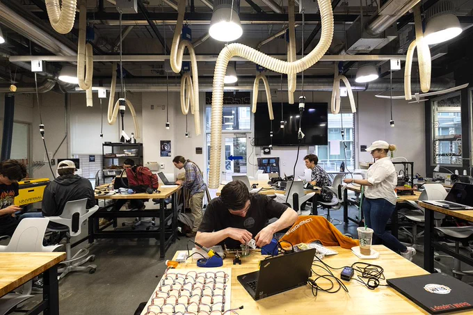 A UNC student works on a project in BeAM. Large ventilation hoses hang from the ceiling. Other UNC students work at tables in the background.