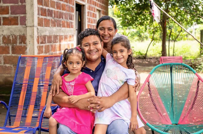 Family of four smiling and sitting together outdoors, with colorful chairs nearby.