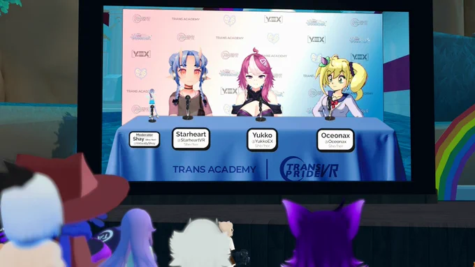 A virtual panel is taking place inside a VRChat instance, shown on a large in-world screen. Three VTuber panelists - Starheart, Yukko, and Oceonax - are seated at a table with a blue cloth labeled “TRANS ACADEMY” and “TRANS PRIDE VR.” Each has a name tag displaying their handle and pronouns (she/her). The panel is moderated by Shay, whose name tag is on the far left. Behind them is a backdrop with various logos. In the foreground, various VRChat avatars are watching the panel, and a rainbow stand to the side of the stage.