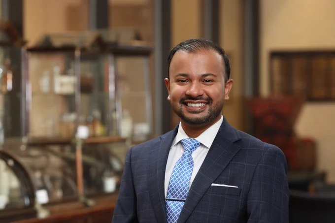 A professional headshot of Justin Samuel, Ph.D. He is smiling brightly to camera in a blue suit and tie.