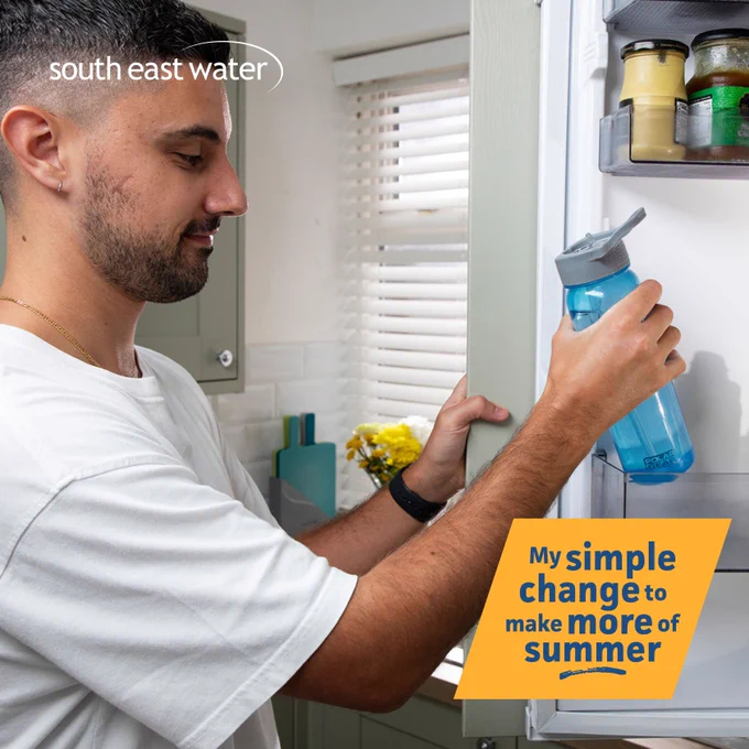 Close up photo of a man wearing a white t-shirt, smiling while putting a blue reusable water bottle filled with water, in a fridge door. The South East Water logo is in the top left hand corner, with  the words 'My simple change to make more of summer' on an orange box, in the bottom right hand corner.