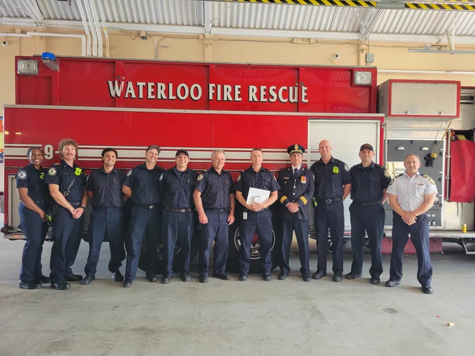 Captain Ryan Kit stands with fellow Waterloo Fire Rescue members in front of a red fire truck. The group is smiling in celebration of Captain Kit receiving his 20-year Federal Medal for dedicated service.