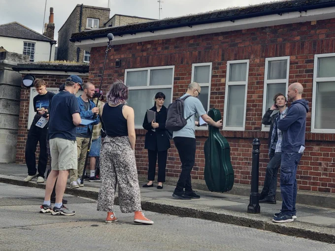 Cast and crew preparing for a shot of a cello case and two leads outside a building.