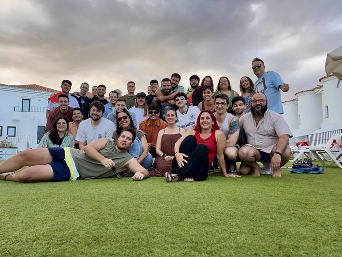 En la foto unas 35 personas (leanminders) sonrientes sentadas sobre un césped verde, mirando a la cámara. Al fondo hay bungalós blancos, varias hamacas y se intuye la presencia de una piscina. El cielo está ligeramente nublado, pero el ambiente transmite alegría, cercanía y diversión.