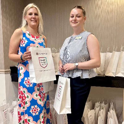 Two Cambridge International Education colleagues getting ready to welcome delegates to the Cambridge Schools Conference 2025 in Florida. They are holding promotional event tote bags, standing in front of a table with more bags and event materials.