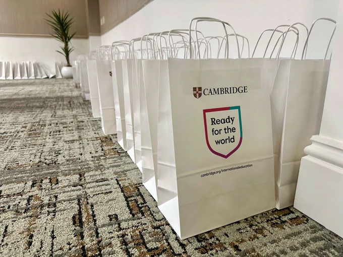 A row of white Cambridge International Education branded bags with "Ready for the World" printed on them, lined up on a carpeted floor.