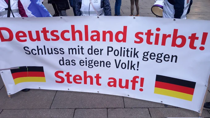 Frontbanner mit der Aufschrift: 
Deutschland stirbt! (fett, in roter Schrift)

Schluss mit der Politik gegen das eigene Volk! (in schwarzer Schrift)

Steht auf! (fett, in roter Schrift, links und rechts ist eine Schlandfahne aufgedruckt)
