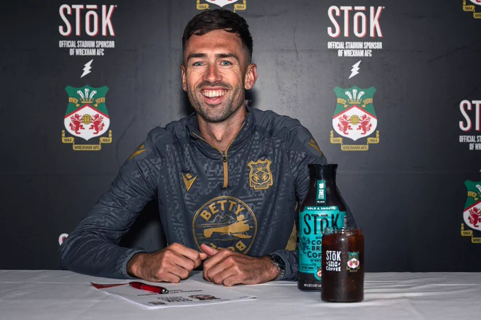 A photo of Ryan Hardie smiling after joining Wrexham AFC on a deal until 2028 - a bottle and a glass of STōK Cold Brew Coffee are on a table in front of him. 